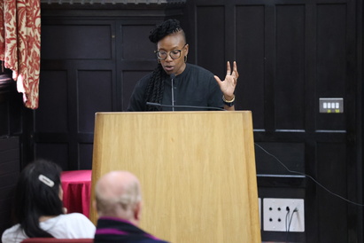 Professor Tendayi Achiume giving a lecture at a podium, in the lecture hall of The Divinity School, St John's College. She is standing in front of a wood panelled wall, facing outwards towards her audience. 