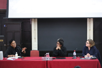 The table at the Alfred Dubs Lecture with Professor Tendayi Achiume on the left, Professor Vasuki Nesiah in the centre, and Dr Tugba Basaran on the right. The table has red covering, microphones at each chair, a carafe of water and glasses in the centre.